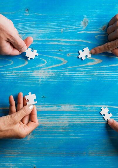 Four people's hands assembling white puzzle pieces on a blue wooden table, symbolizing conflict resolution and collaboration in event planning.