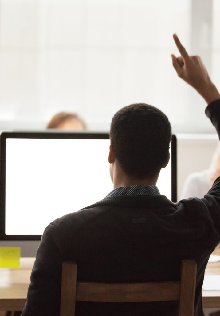 Rear view of a man raising his hand in a meeting, with a blank computer screen and coworkers in the background.