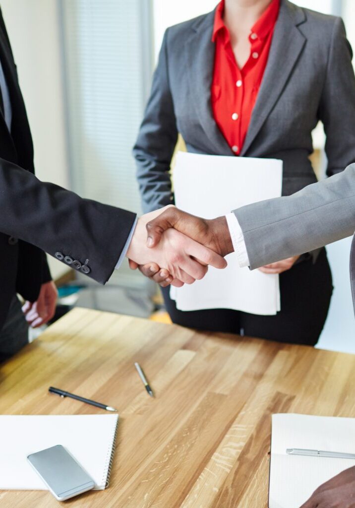 Two business professionals shaking hands across a table in an office setting, with documents and smartphones on the table.